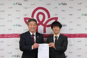 丹波市と書かれたパネルの前で委嘱書を掲げるスーツ姿の市長の林時彦と、地域おこし協力隊の滝沢仁悟の写真
