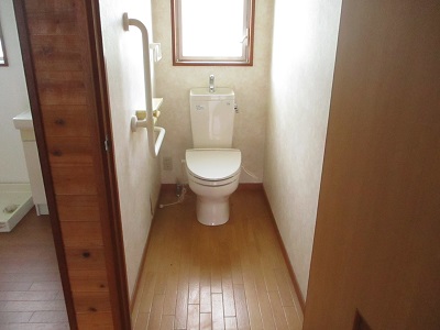 白を基調としたトイレで、床はフローリング、右手側に白い手すりがある写真