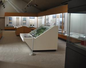 中央に白い展示ケースがあり、奥には茶色の展示ケース、壁面にガラス面の展示スペースがある室内の写真