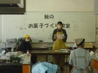 「秋のお菓子作り教室」と書かれたホワイトボードの前で紙を持った女性が立っている写真