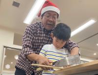 サンタ帽を被った男性と一緒にのこぎりで板を切っている子供の写真
