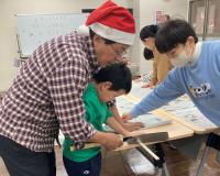サンタ帽を被った男性と子供が一緒に、他の子供が手で抑えている板をのこぎりで切っている写真