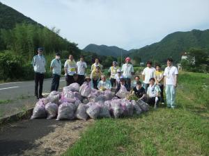 屋外にて作業着を着た複数人の人が並び、手前にはゴミ袋が山積みになっている写真