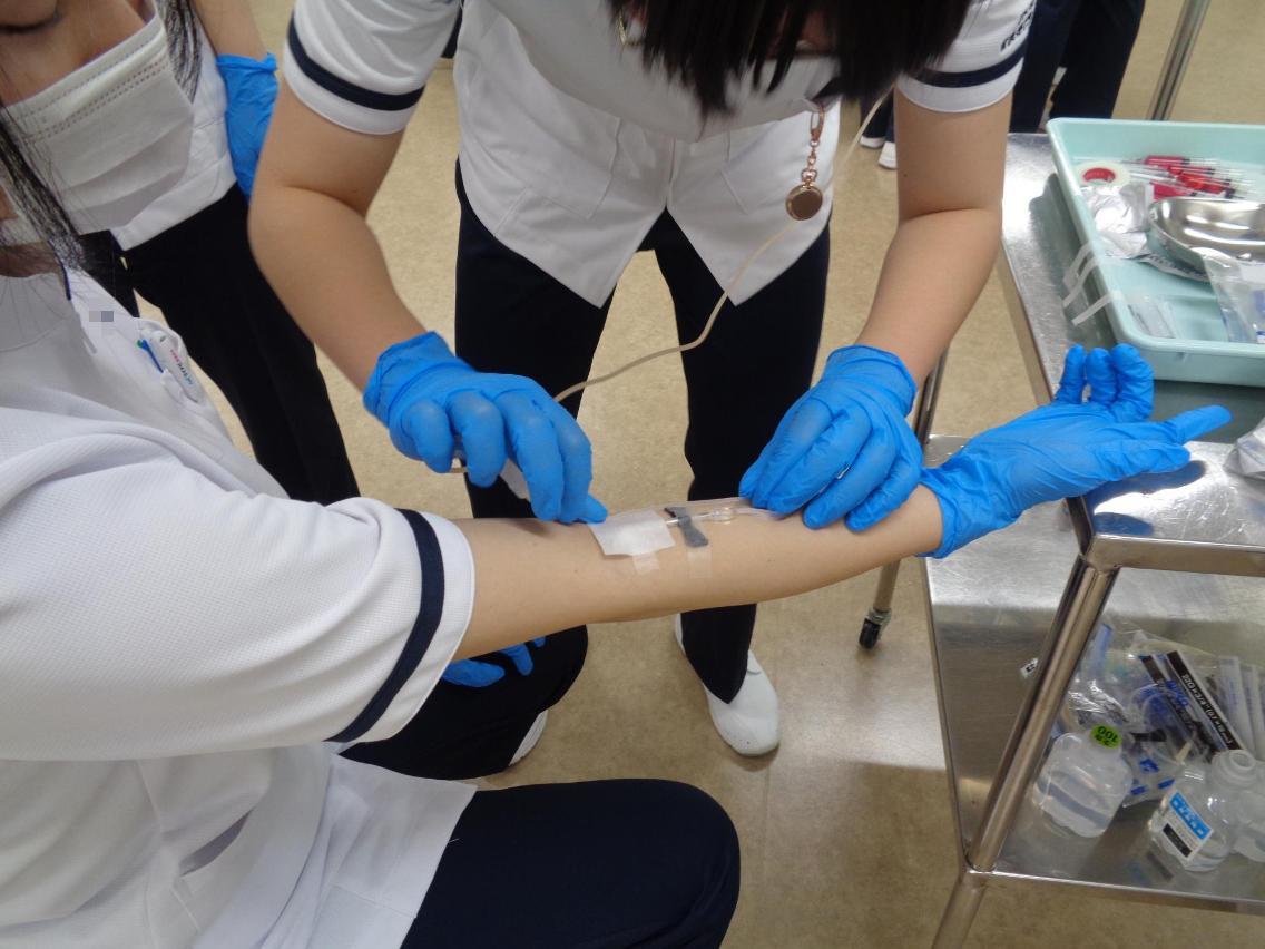 患者役の学生の腕に点滴針の位置を決めて、テープで固定している看護学生の写真