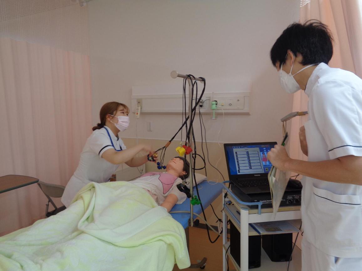寝ているシミュレーターの胸元に診療器具を当てている看護学生と、見守っている教員の写真