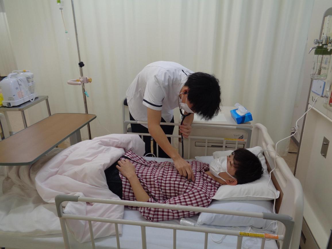 ベッドに寝ている患者役の学生の胸元に、聴診器を当てている看護学生の写真