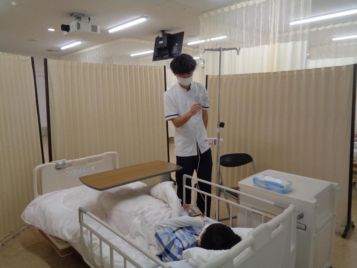 ベットに寝ている患者役の学生の横で、点滴の速さを調整している看護学生の写真