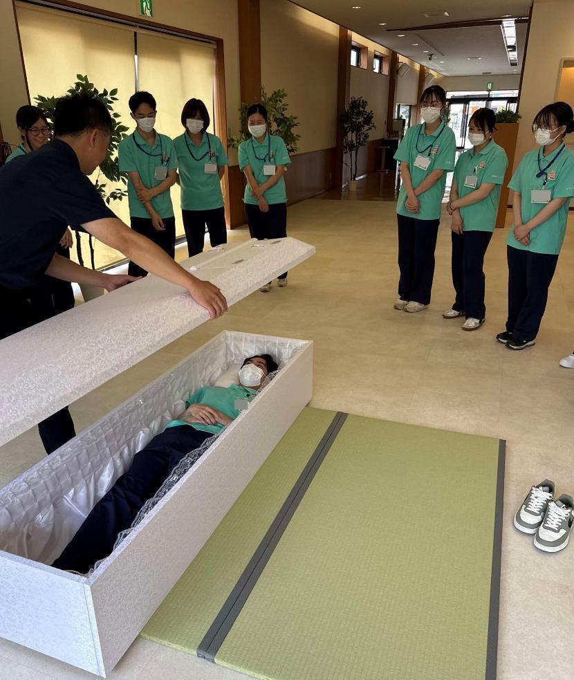 葬祭ディレクターが、学生が中に入った棺のふたを、かぶせようとしているところの写真