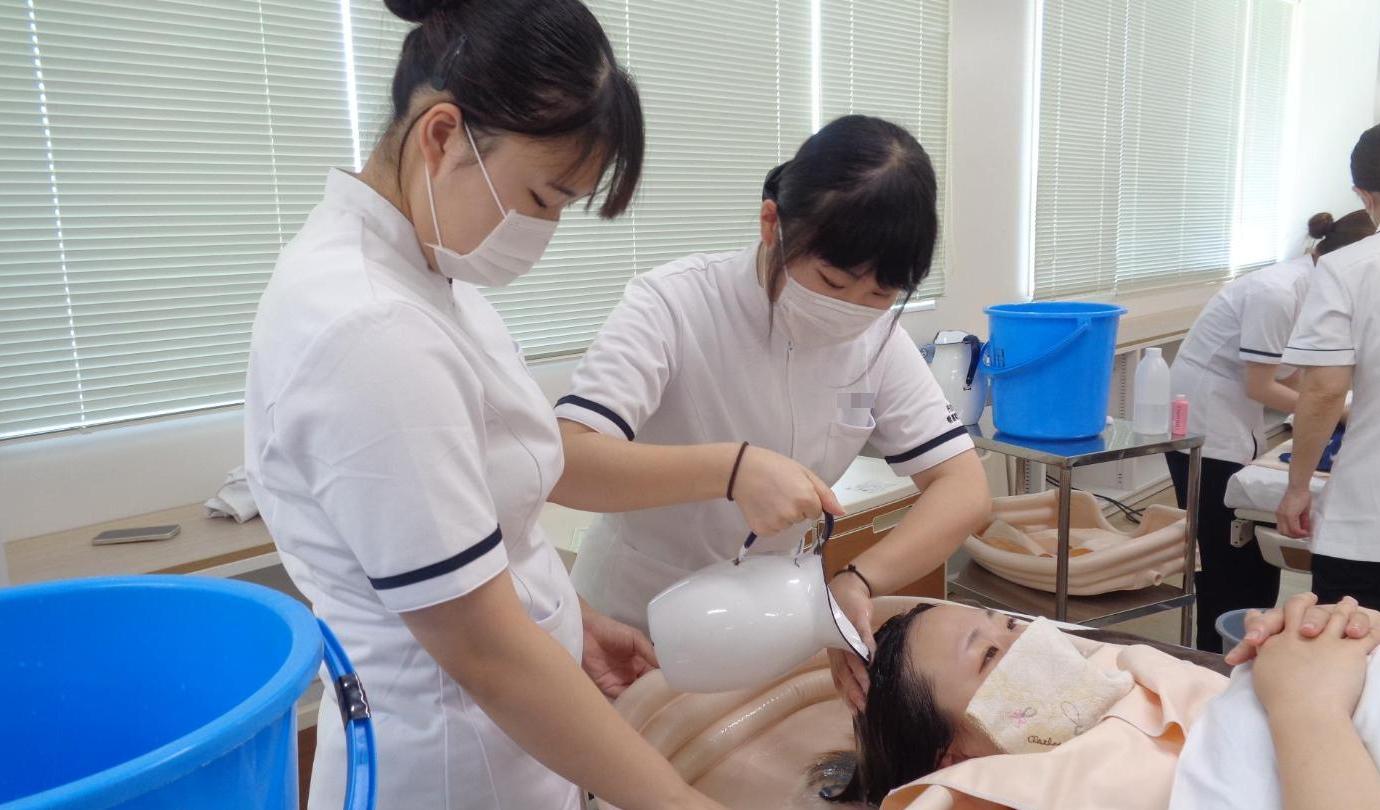 学生がベッド上で学生の洗髪をした後、ピッチャーの湯をかけて髪をすすいでいるところの写真