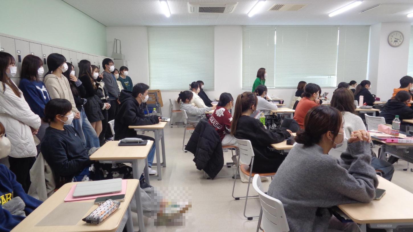 卒業生たちが教室の後ろに立って、後輩の学生たちの様子を見ている写真