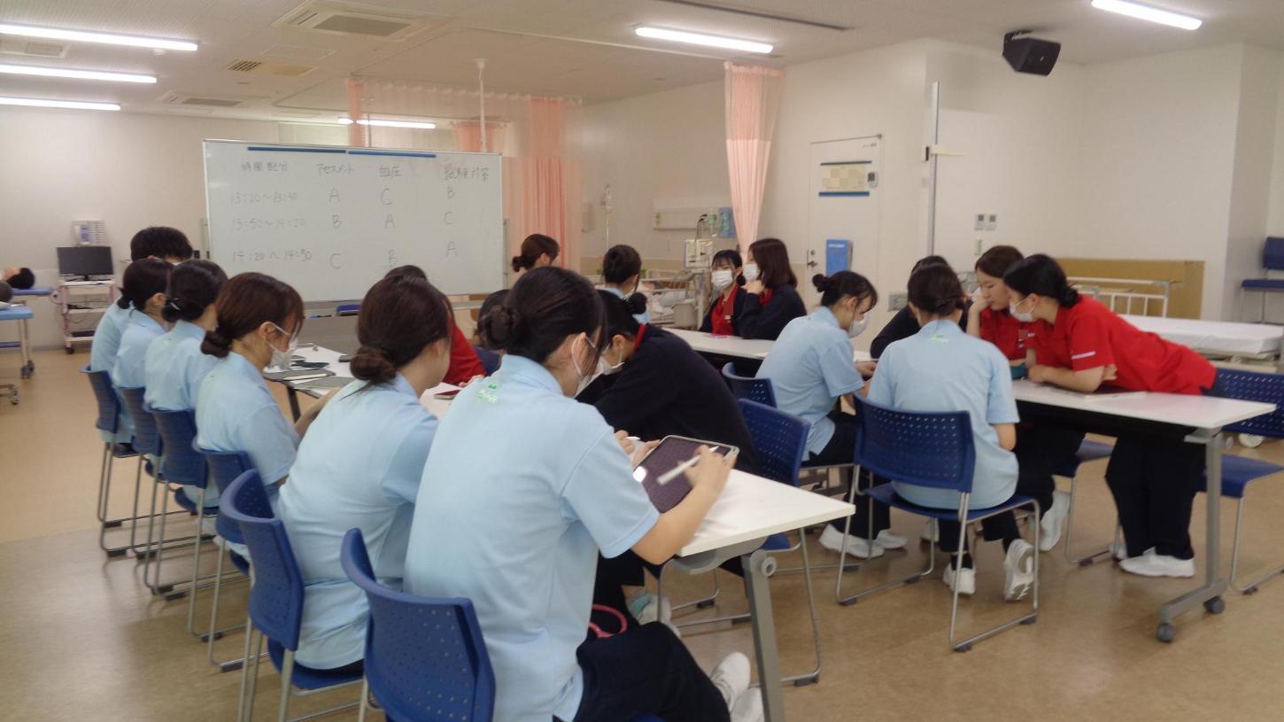 実習室で、約10人ずつが2列の机に向かい合って座り、学生がアセスメントに関する意見交換をしているところの写真