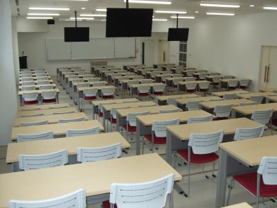 前面にホワイトボードと三つのモニターがある、階段状に机と椅子が並べられた広い教室の写真