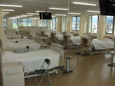 看護ベッドが一定の間隔を開けて並べられている、広い部屋の写真