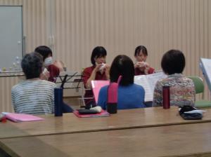 えんじ色のTシャツを着た女性3人がオカリナを吹き、年配の女性3人が楽譜を手に持っている写真