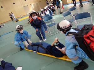 ヘルメットとマスクをつけたつなぎ姿の男性が担架に人を乗せて救助訓練をしている写真