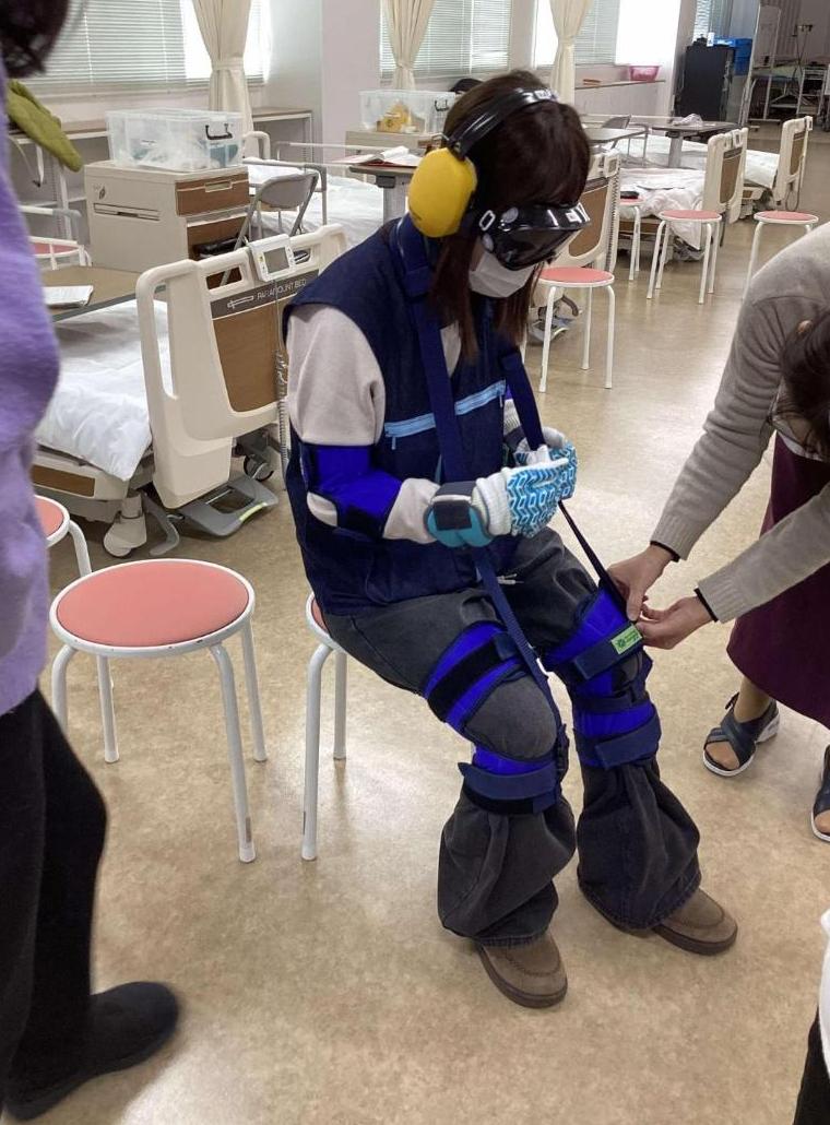 老化体験スーツを着た学生のひざパッドをベルトで調整しているところの写真