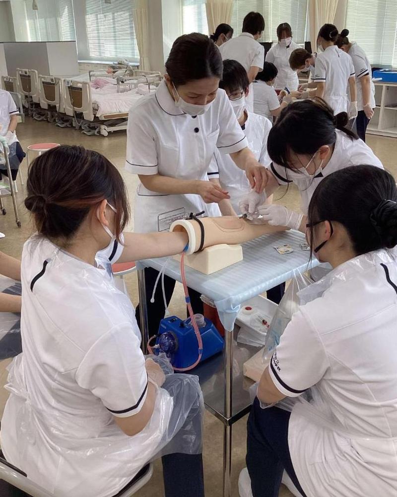 患者役の腕に固定した上腕モデルに採血をしている学生と見守る教員と学生たちの写真