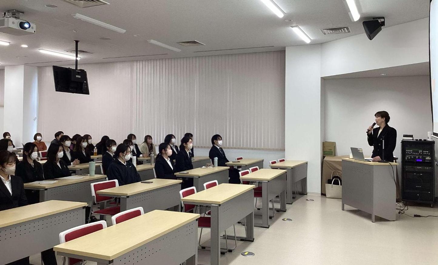 スクリーンの前でマイクを持った講師が、大勢の看護学生を前に講演している様子の写真