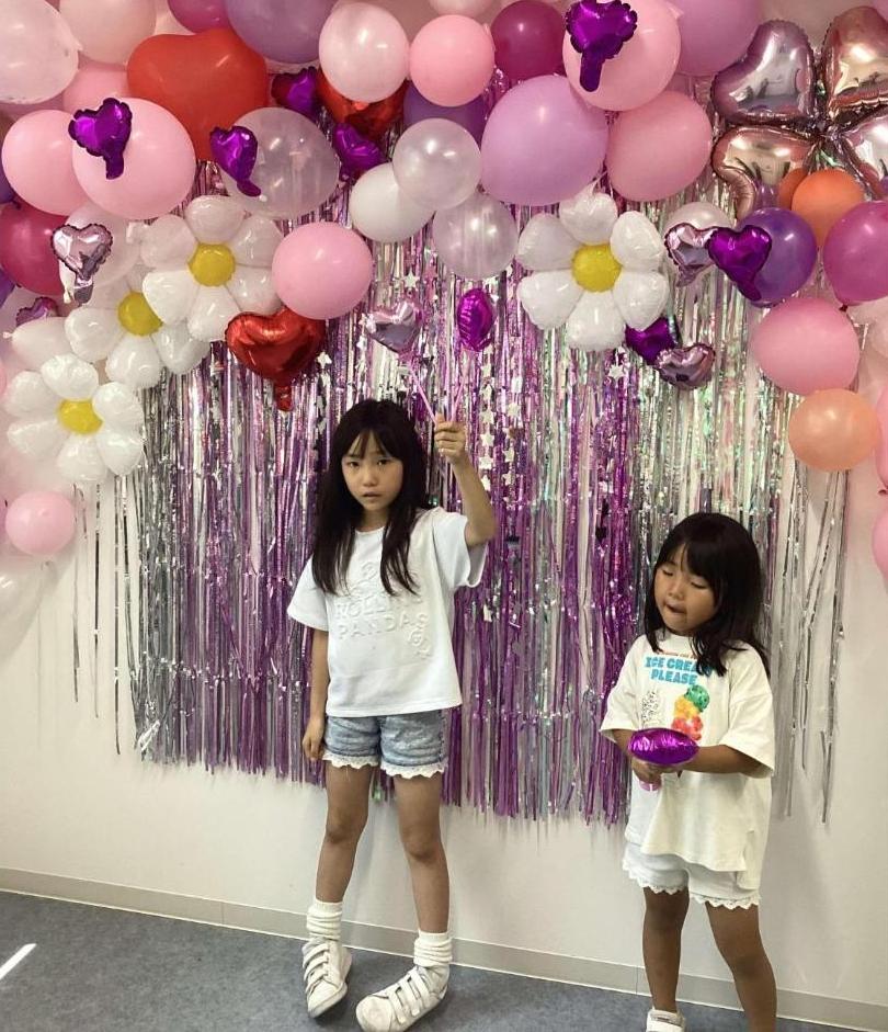 参加者2人の子どもさんが、ピンクや赤や紫など可愛い色の風船を使って学生達が作ったフォトスポットの前に立っているところの写真