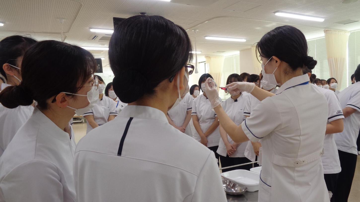 教員の注射器に薬を吸引するデモンストレーションを看護学生たちが見ているところの写真