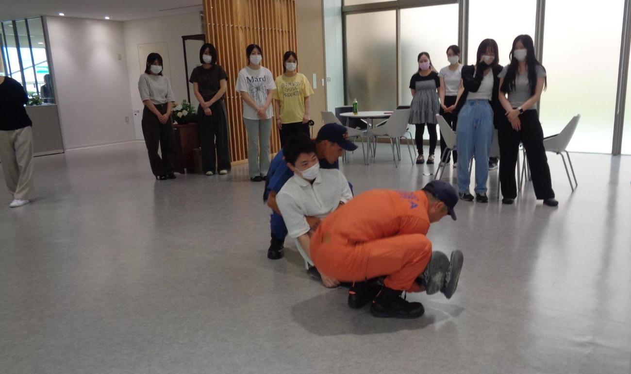 消防署職員が2人で負傷者を搬送する方法を説明し、それらを見ている学生たちの写真