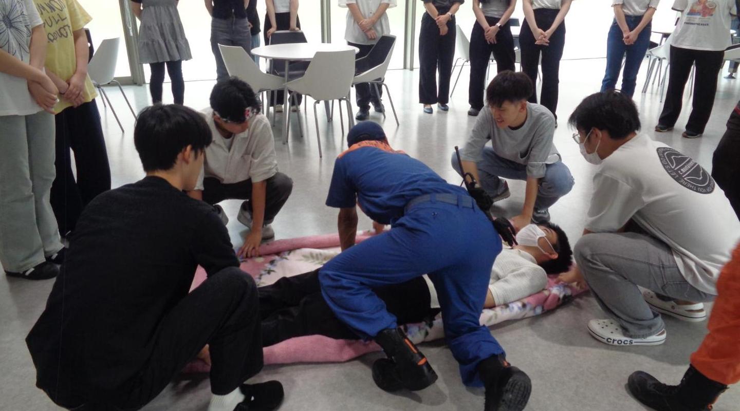 消防署職員の説明を聞いて負傷者の下に毛布を敷き、毛布を丸めて持ち、4人で搬送しようとしている学生たちの写真
