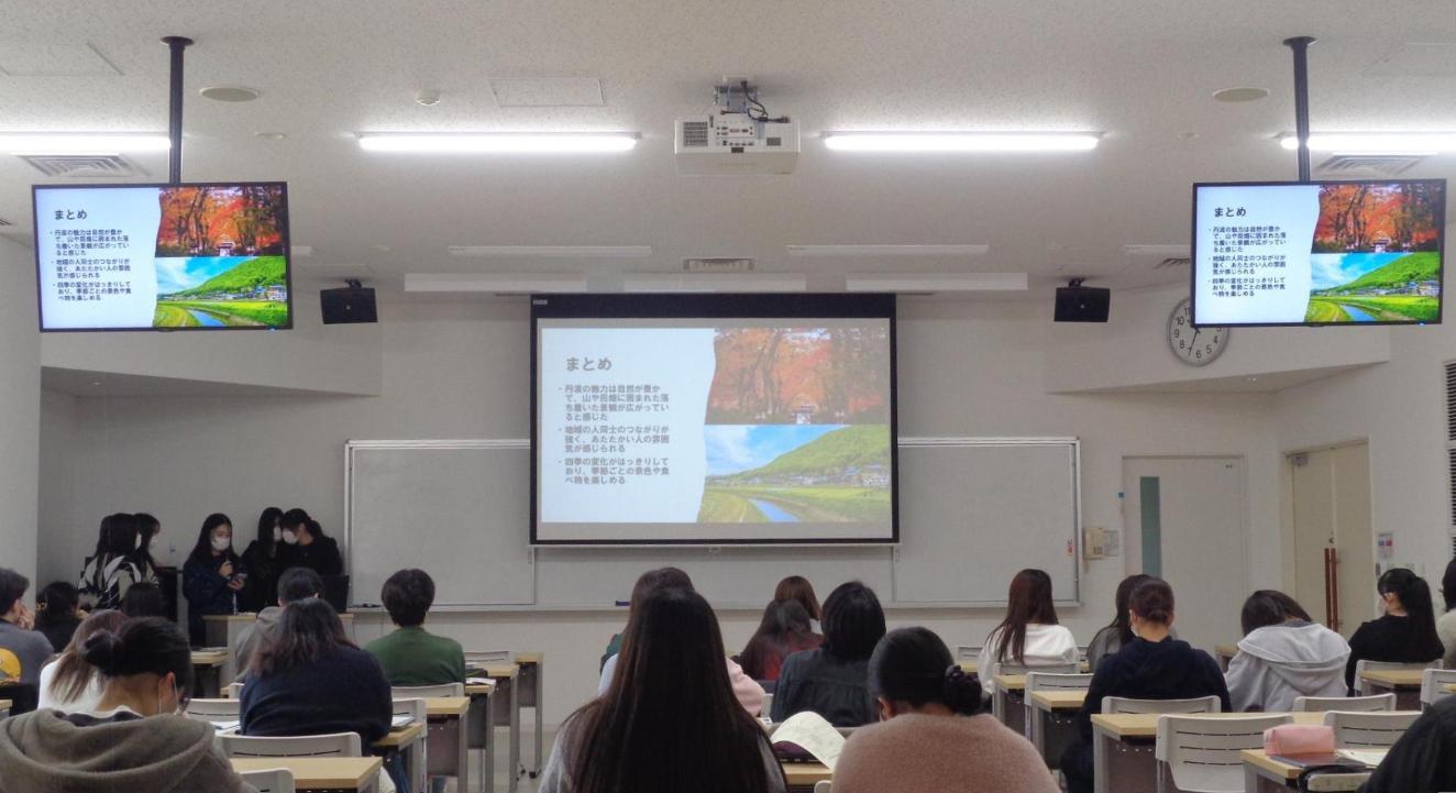 スクリーンに紅葉と新緑の写真が映され、マイクのある檀の前で数人の学生が並んでまとめを話している写真