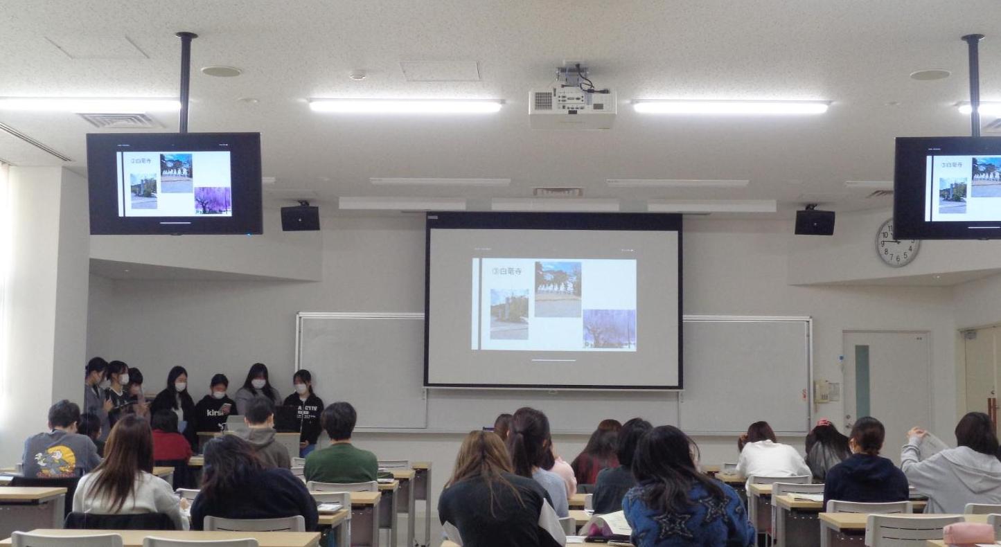 校内発表、スクリーンに市島町白毫寺の藤の花・七福神の石像などの写真が映され、大勢の学生がそれを見ている写真