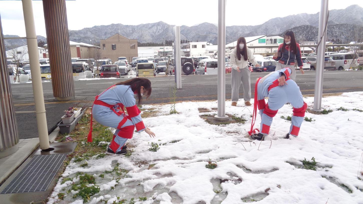 掲揚柱のそばで、雪を触るのに前かがみになろうとしている老化体験スーツを着た学生たちの写真