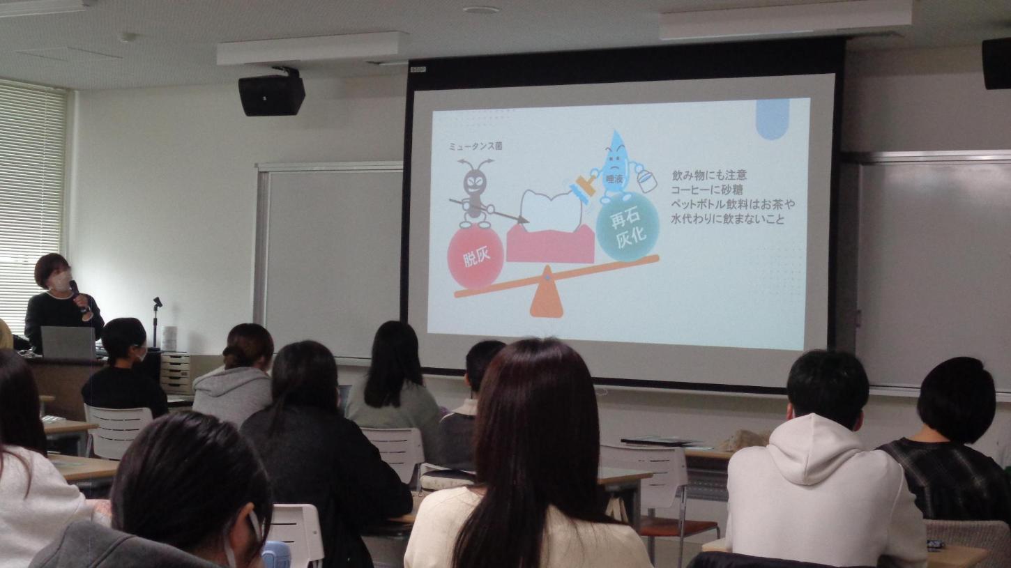 歯に悪さするバイキンと再石灰化をする働きのキャラクターが映されたスクリーンを説明している歯科衛生士と聞いている学生たちの写真