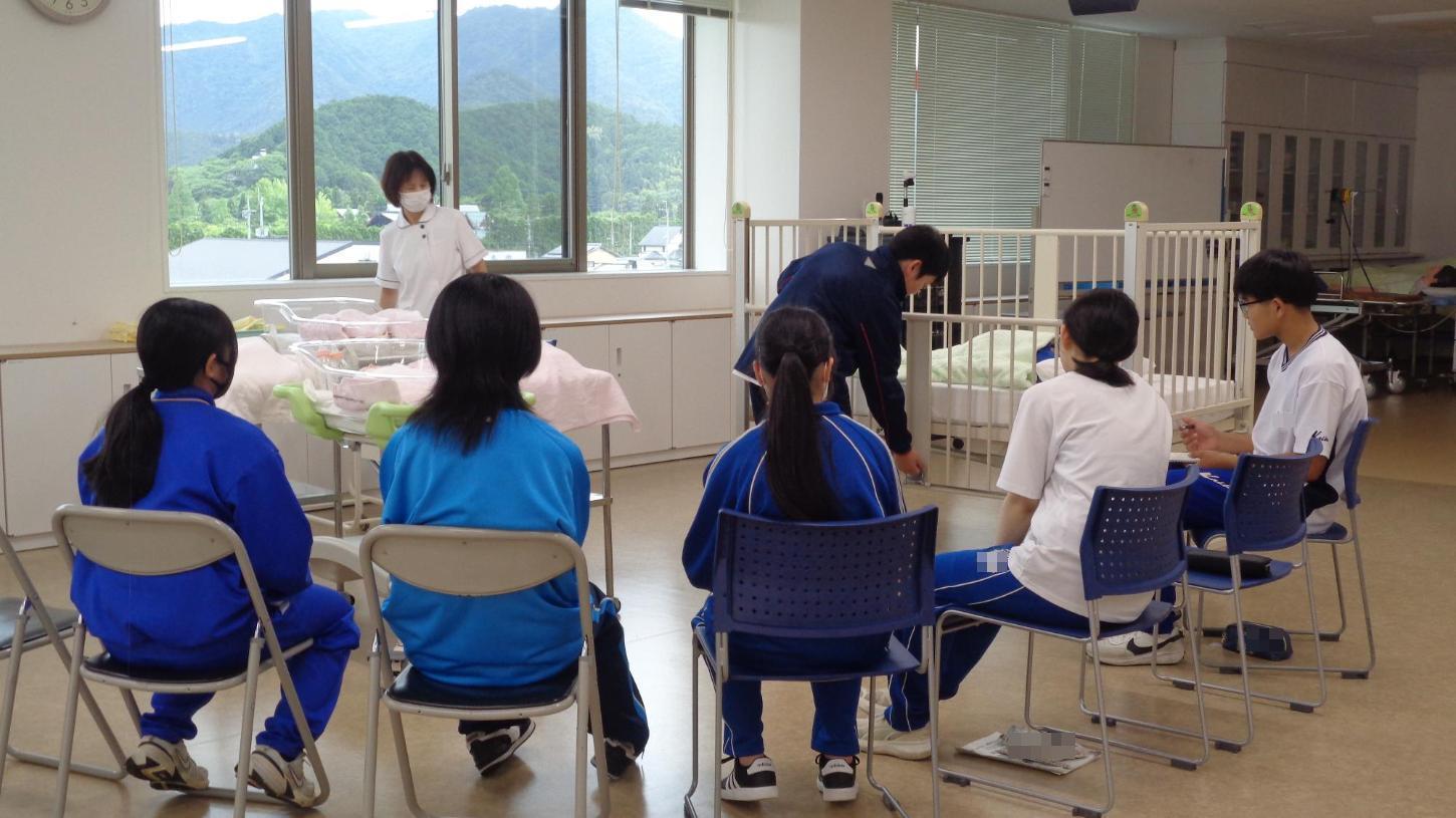 ベビーベッドの使い方の説明を聞き、柵を両手で上げ下げしている中学生とそれをみている中学生達の写真