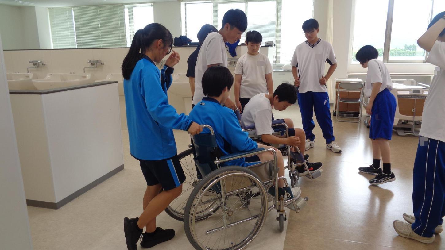 車椅子2台にそれぞれ乗り、介助者が後ろに立ち、レバーを確認している中学生を見ている周りの中学生達の写真