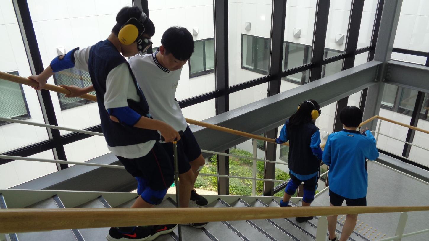 老人体験スーツを着た中学生と横に付き添いの中学生が階段を下りているところの写真