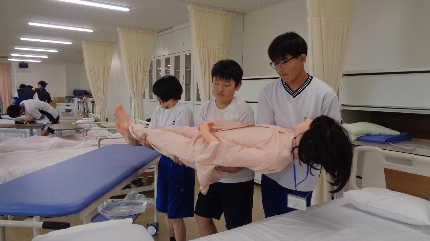 患者役人形を息を合わせてベッドからストレッチャーへ移動している中学生達の写真