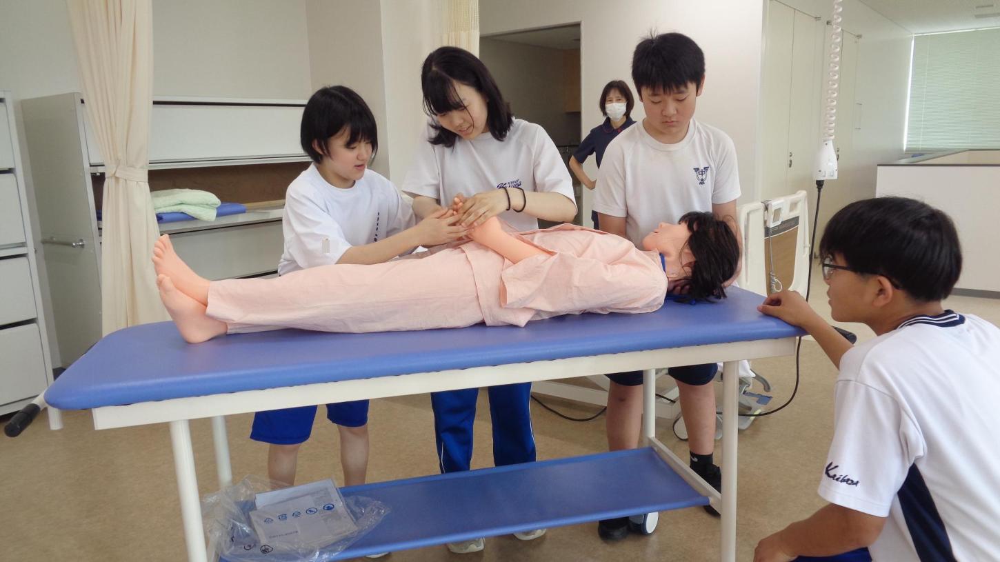 ストレッチャーからベッドに移動する前に患者役人形の手を組ませようとしている中学生達の写真