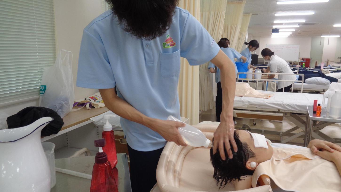 学生がベッド上で学生の洗髪をした後、ボトルシャワーの湯をかけて髪をすすいでいるところの写真