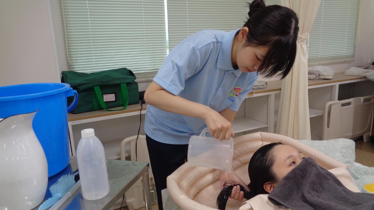 学生がベッド上で学生の洗髪をした後、ピッチャーの湯をかけて髪をすすいでいるところの写真