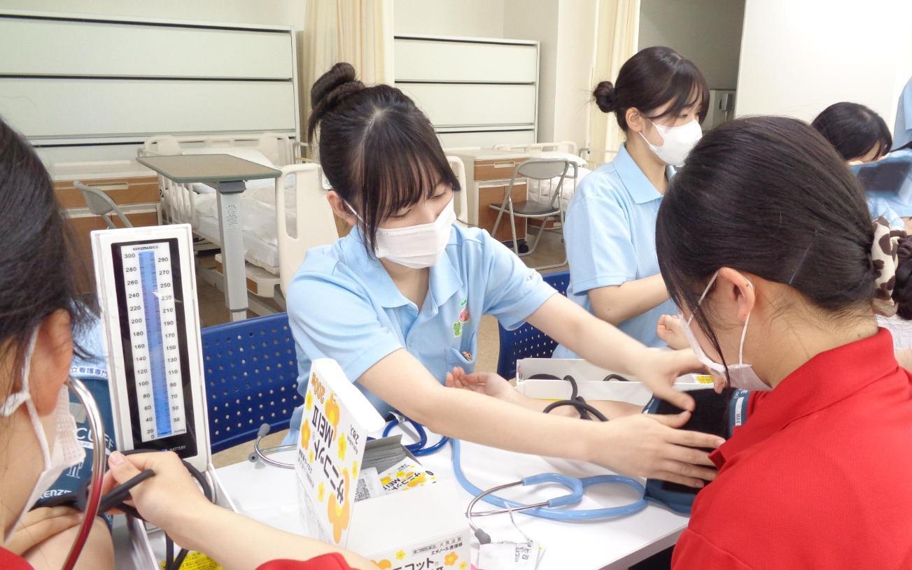 実習室で、数人が机に向かい合って座り、学生が先輩の腕に医療器具を巻いて、血圧を測定し始めようとしているところの写真