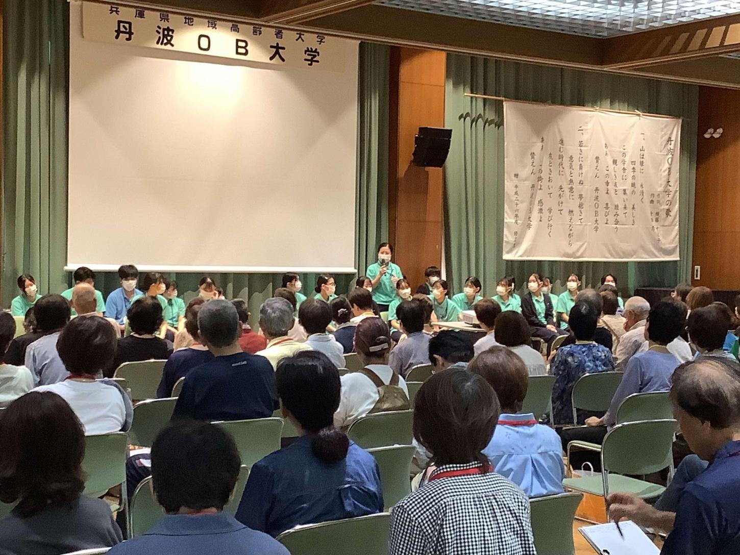 緑色のTシャツを着た学生たちが2列に並べられた椅子に座って挨拶をしている写真