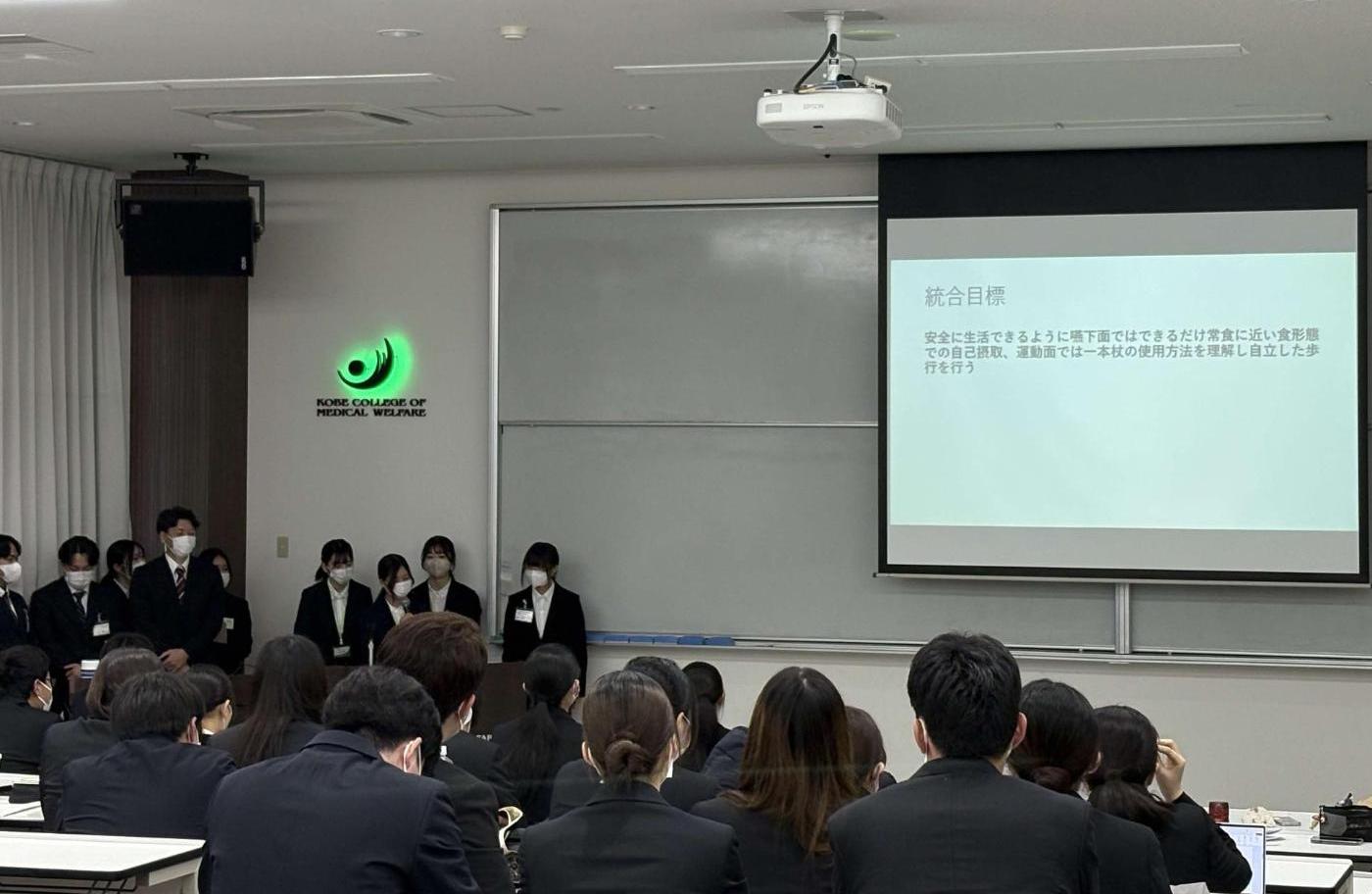 スクリーンに投影された資料を説明しているグループの学生たちと、机に座って資料を見ている学生たちの写真