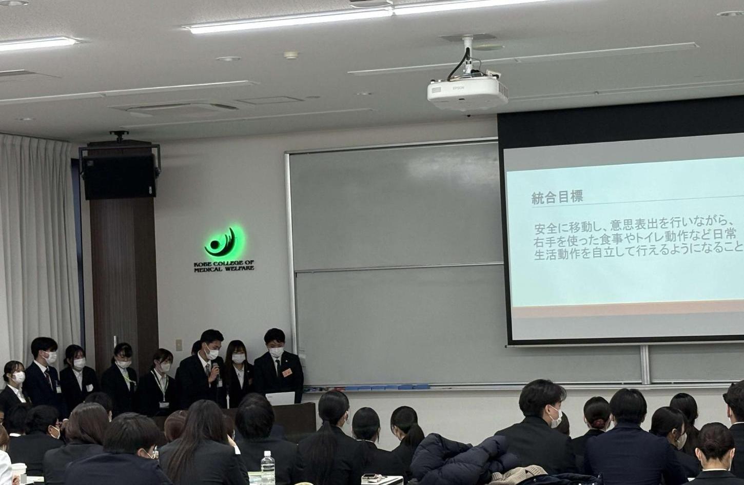 スクリーンに投影された資料を説明しているグループの学生たちと、机に座って資料を見ている学生たちの写真