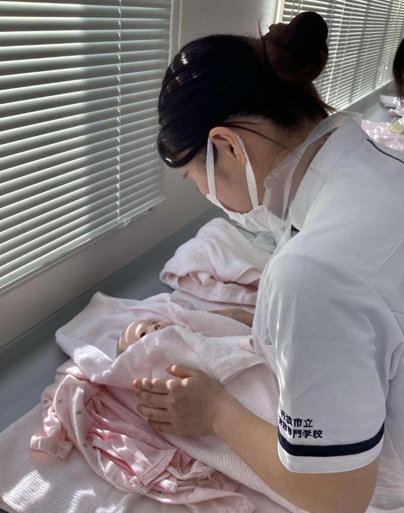 台の上で、沐浴が終わった新生児人形を、タオルに手を添えてやさしく包んでいる学生の写真