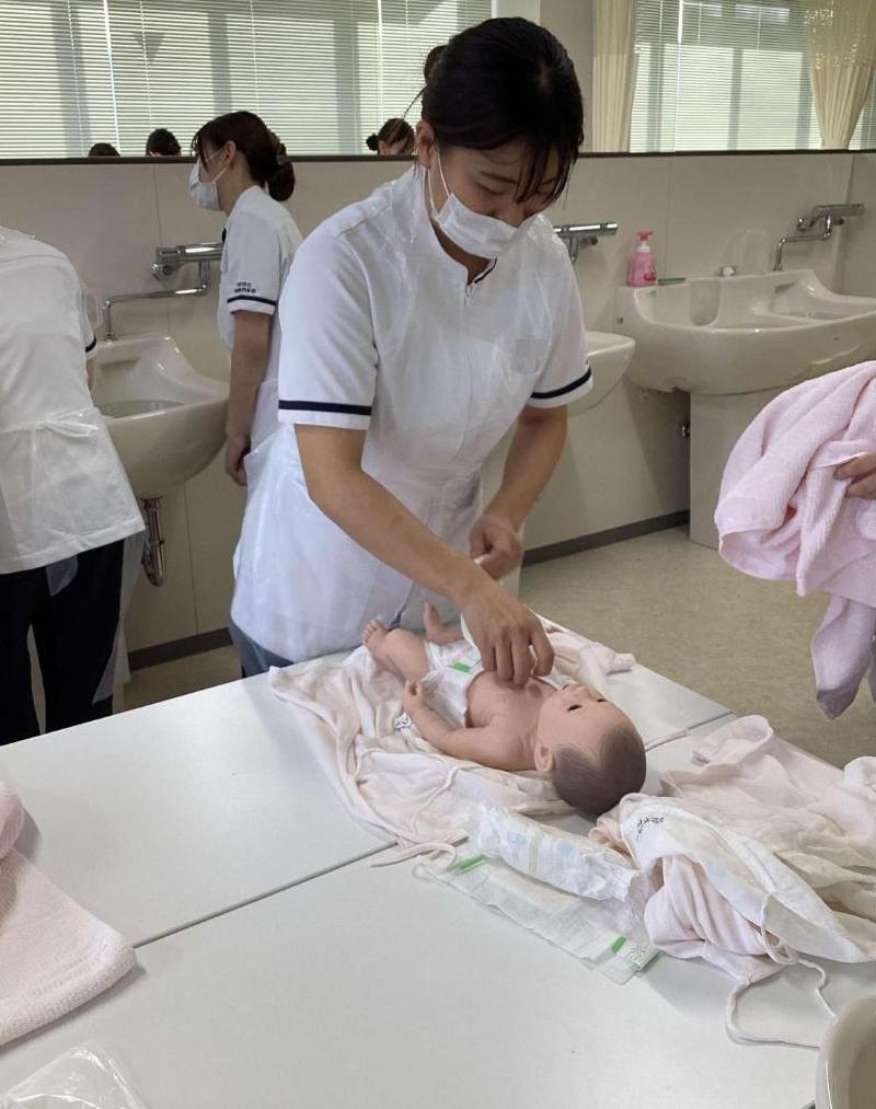 台の上で沐浴が終わった新生児人形に肌着を着せている学生の写真