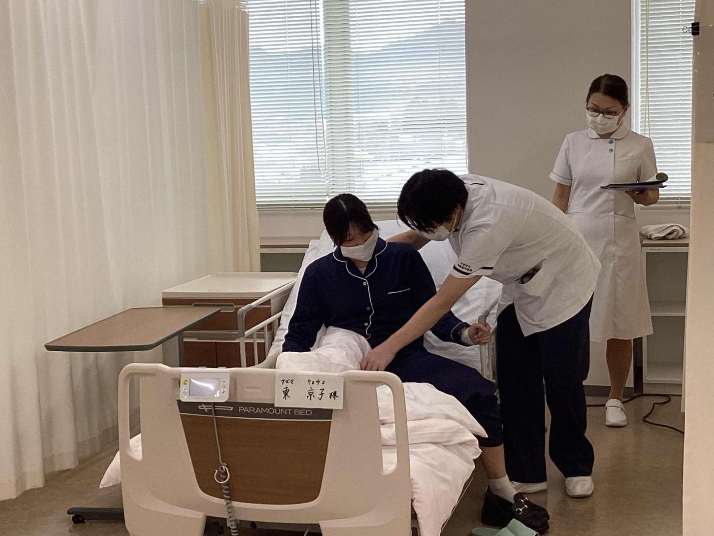 患者がベッドがら下りるのを介助している学生と、見守っている教員の写真