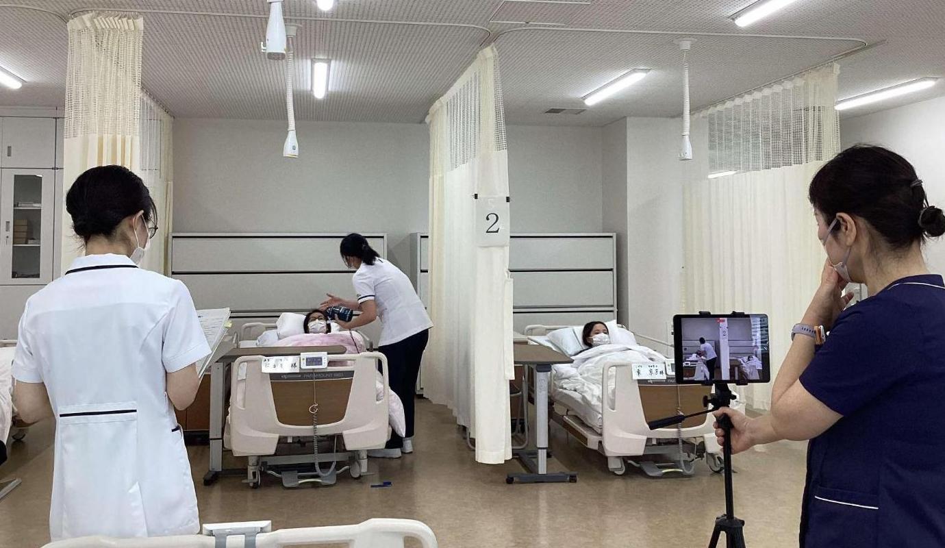 学生が患者を看護しているところを見守っている教員と、撮影をしている教員の写真