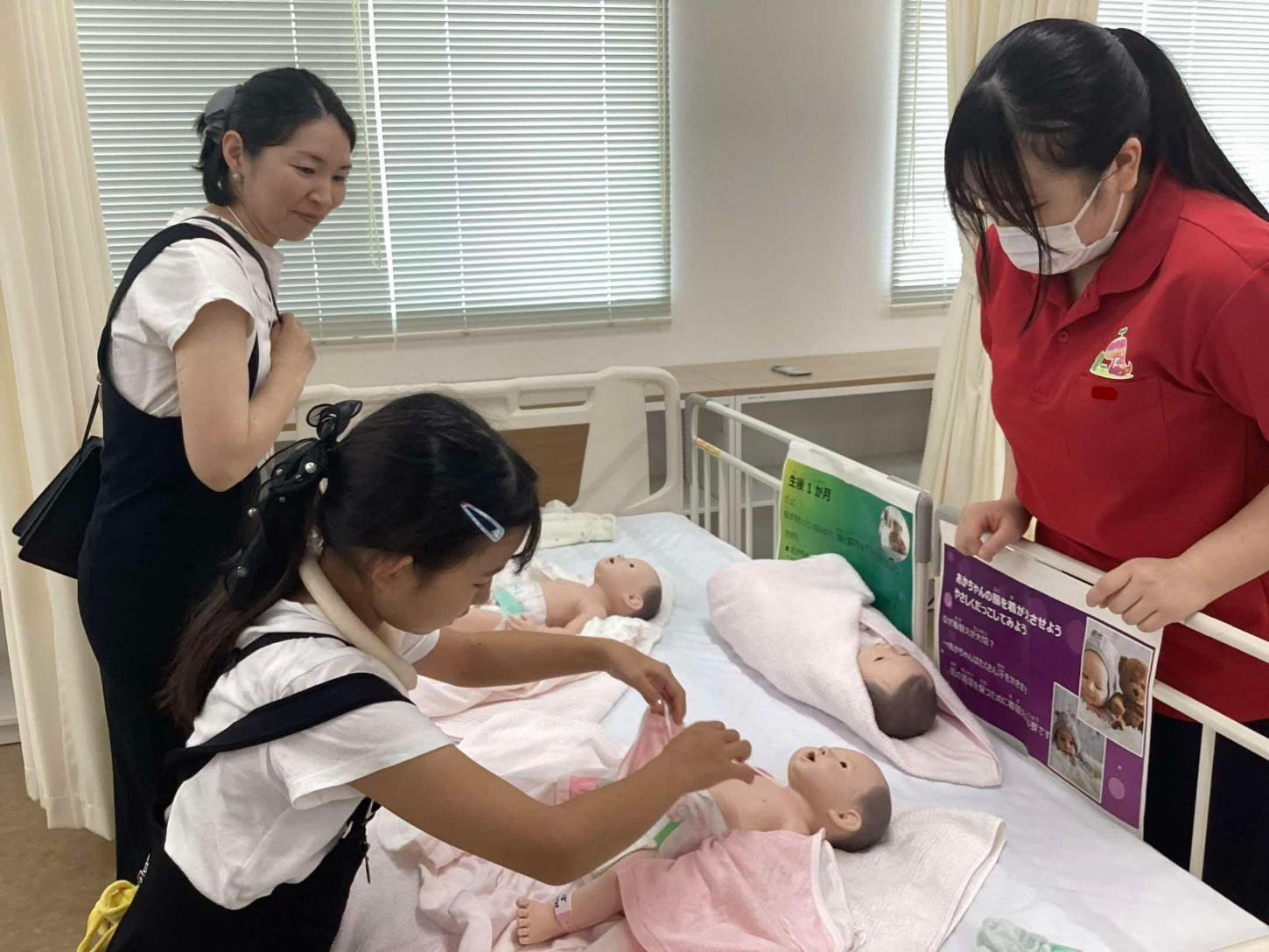 赤ちゃん人形を着替えさせている小学生と見守っているお母様と学生の写真