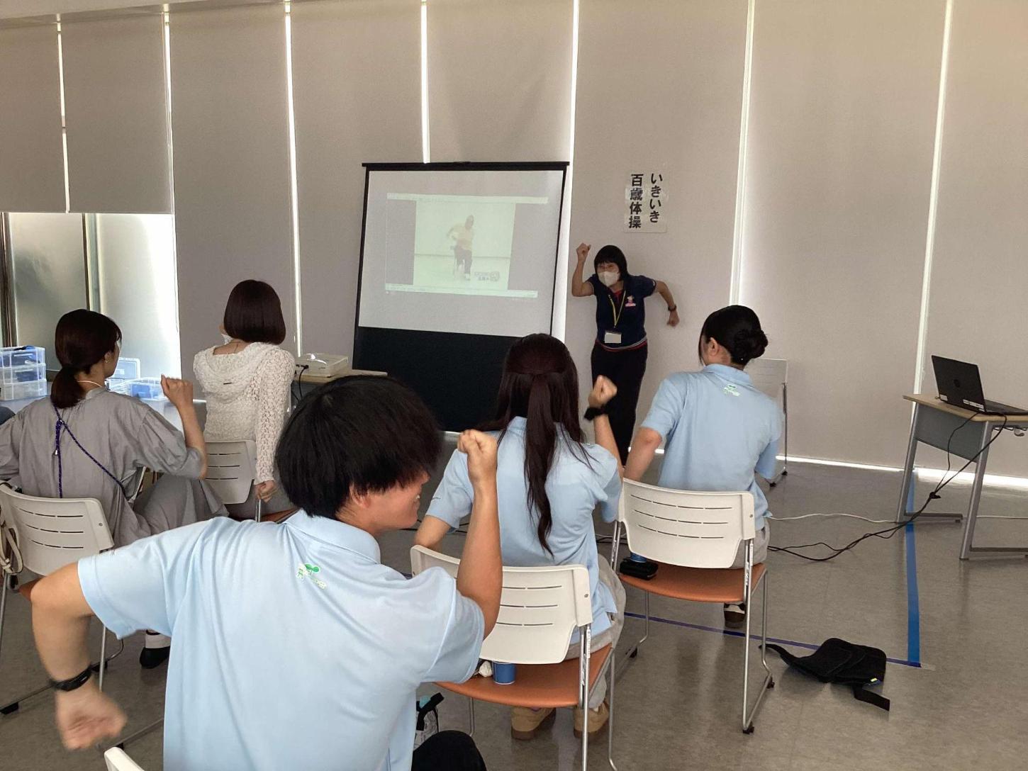 講師の説明を聞き、椅子に座って「いきいき百歳体操」をしている参加者の方々と学生達の写真