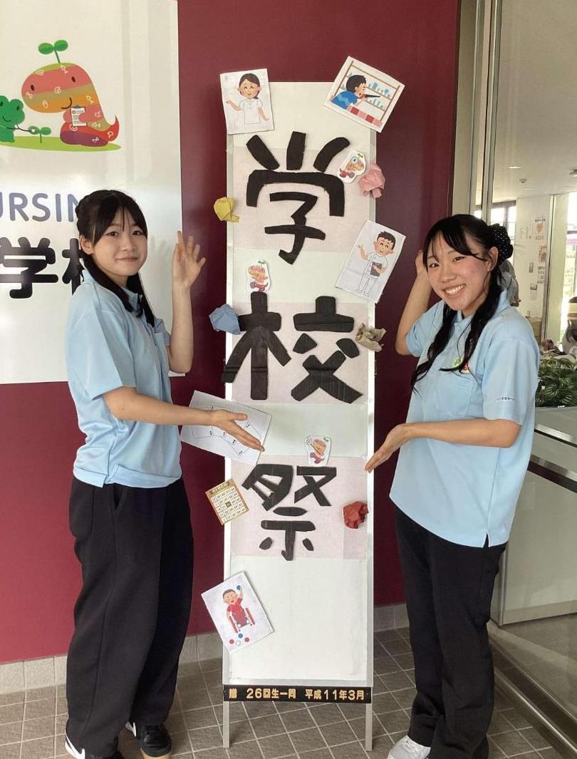 玄関の「学校祭」パネルの横で、学生二人が手を広げて立っているところの写真