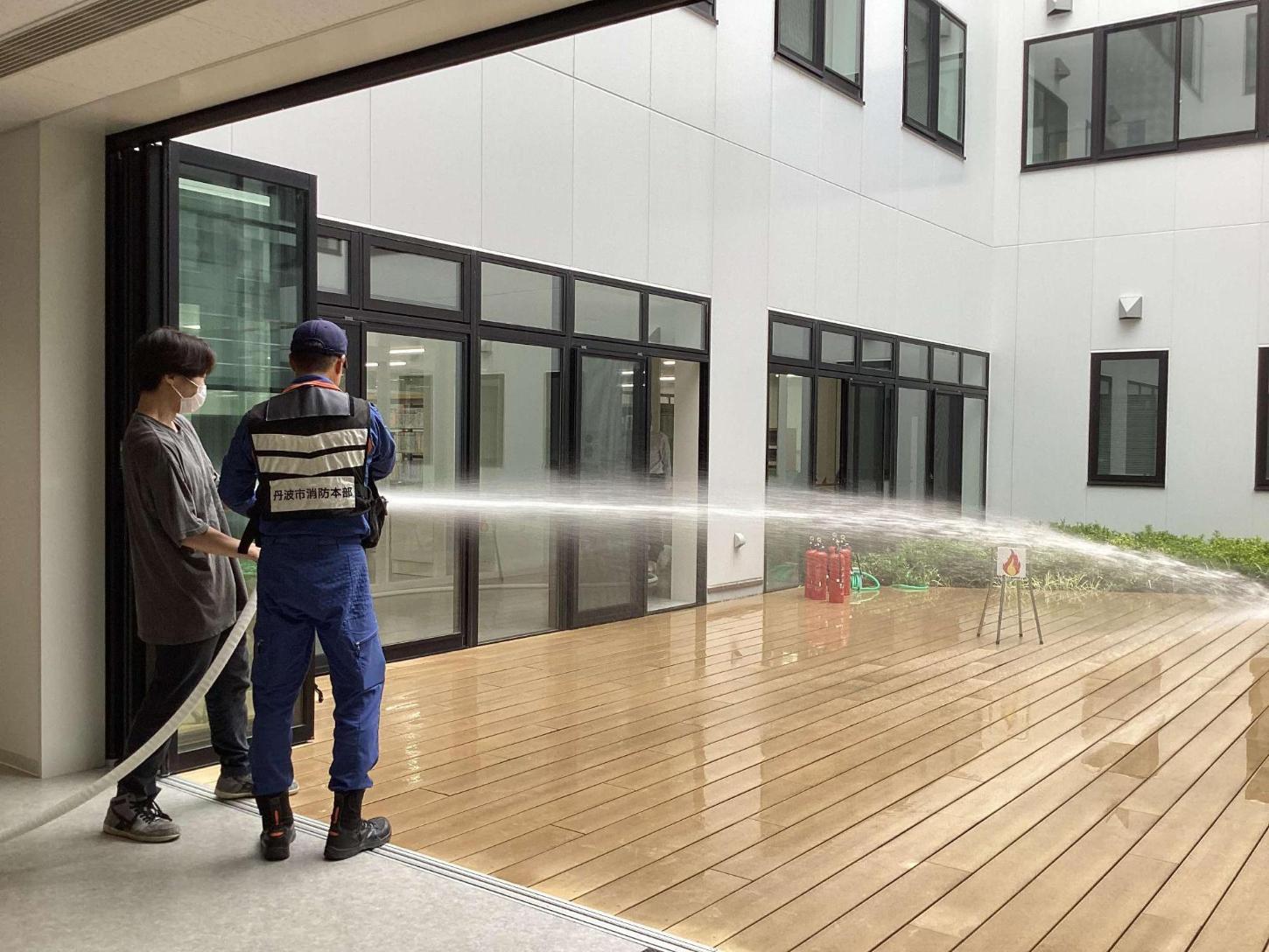 屋内消火栓のホースを消防署員と一緒に持ち、火元の的に向けて放水をしている学生の写真