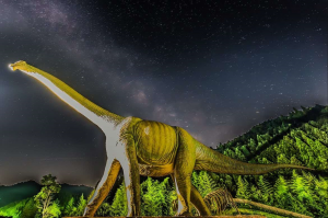 針葉樹の山と星空を背景に歩く、首の長い草食恐竜の模型の写真